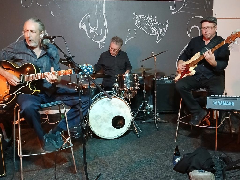 Mojo Webb playing a hollow-body guitar, with drummer and bass player